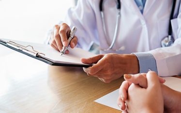 White male doctor holding a pen while pointing at a medical clip board on a wooden table