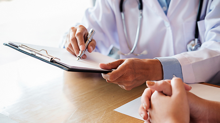 White male doctor holding a pen while pointing at a medical clip board on a wooden table